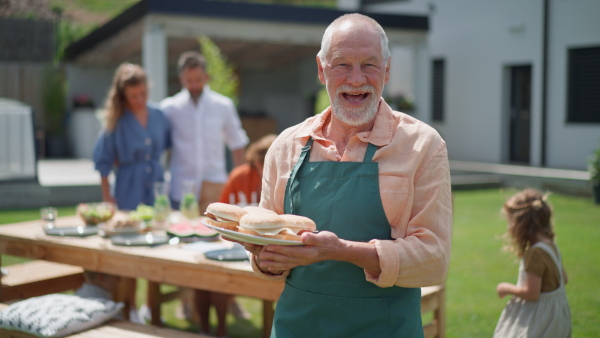 A happy senior man serving burgers at multi generation garden party in summer.