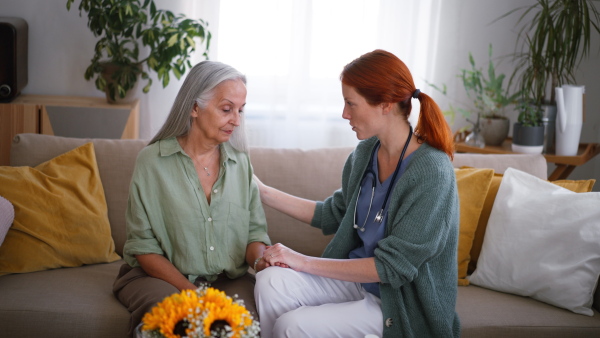 Nurse talking with senior woman, holding her hand and consoling her at her home.