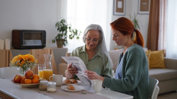 Nurse reading newspaper with senior woman at her home, during the breakfast.