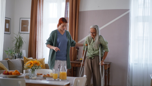 Nurse helping senior woman with walking after leg injury, in her home.