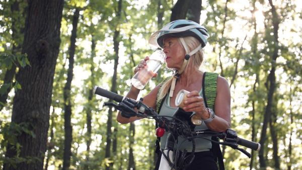 A happy active senior woman biker drinking water from sustainable bottle when sitting on bike outdoors in forest.