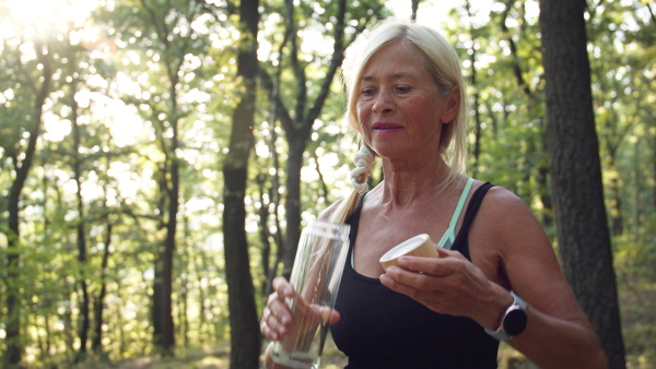 A happy active senior woman drinking water from sustainable bottle when resting in forest.