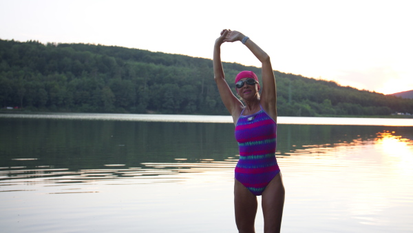 A portrait of active senior woman swimmer standing and stretching outdoors by lake.