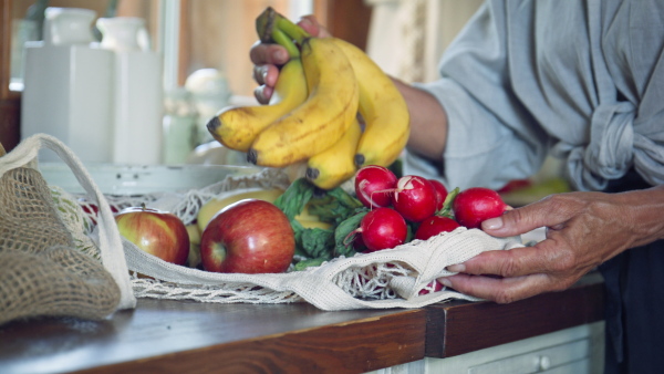 A senior woman unpacking local food in zero waste packaging from bag in kitchen at home.