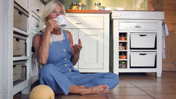 A happy senior woman sitting on floor and drinking tea indoors in kitchen.