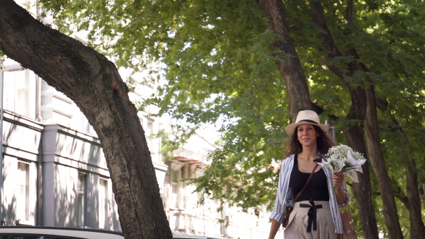 Young woman walking on street under green trees, carrying flowers and greeting. Low angle shot.