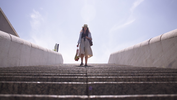 Young woman walking down the stairs outdoors, carrying shopping in zero waste bag on sunny day. Low angle shot.