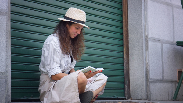 Young woman sitting on street in summer, reading book.