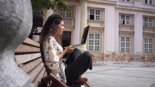 Young woman working on laptop on city bench in historical surroundings with cafe in background.