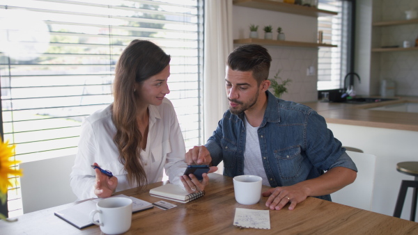 Young couple at home, sitting at the table, using mobile phone for planning of finances.