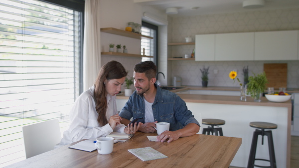 Young couple at home, sitting at table, using mobile phone application for planning of finances.