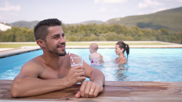 Young man drinking water from a glass at swimming pool while his wife and daughter are playing in the background.