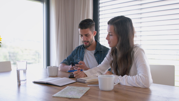 Young couple at home, siting at table, planning travelling, using mobile phone application.