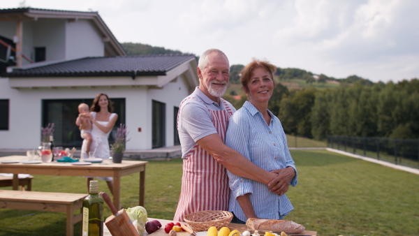 Portrait of happy senior couple in front of modern house, smiling and looking into camera, while preparing barbeque with family.