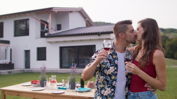 Happy young couple standing in garden in front of new home, kissing and celebrating with wine. Table set for garden dinner party.