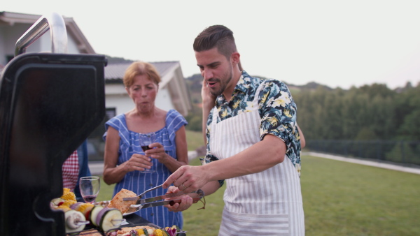 Man preparing food on grill, with his family and friends. Modern house in background.