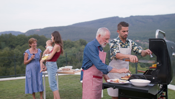 Senior father and adult son preparing food on barbecue. Family standing in background. Beautiful natural scenery.