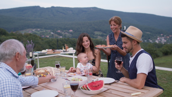 Three generation family having a garden dinner party together, sitting at table. Beautiful natural scenery in background.