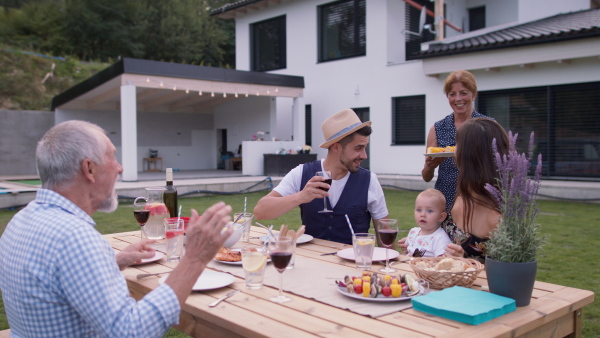 Three generation family having a garden dinner party together, sitting at table. Modern family house in background.