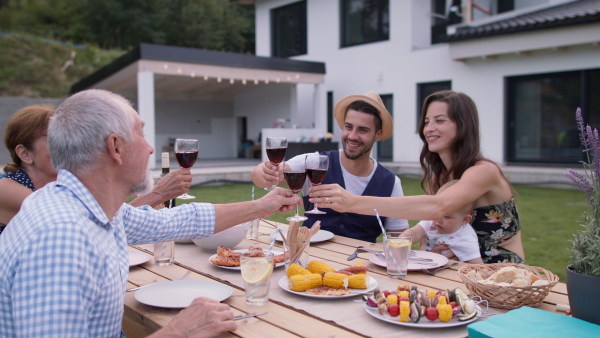 Three generation family having a summer garden dinner party in front of modern family house, making a toast with red wine.