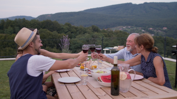 Three generation family having a garden dinner party together, making a toast with red wine. Beautiful nature scenery in background.