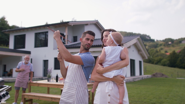 Happy mother, father and baby girl standing in front of house, eating corn knob at barbeque garden party.