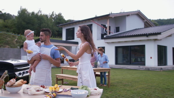 Happy mother, father and baby girl standing in front of house, eating corn knob at barbeque garden party.