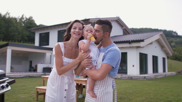 Happy mother, father and baby girl standing in front of house, eating corn knob at barbeque garden party.