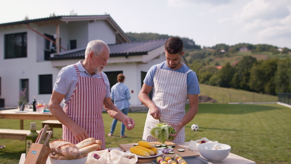 Senior father and adult son standing in garden preparing vegetables for barbecue, modern house in background.