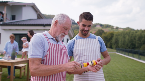 Senior father and adult son standing in garden preparing vegetables for barbecue, modern house in background.