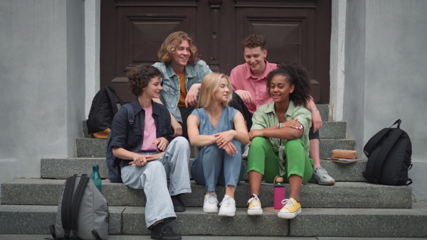 A group of happy teenagers together in front of school, sitting and talking, back to school concept.