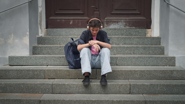 A sad teenager student sitting on stairs in front of school, listening to music. Bullying, discrimination or stress concept.