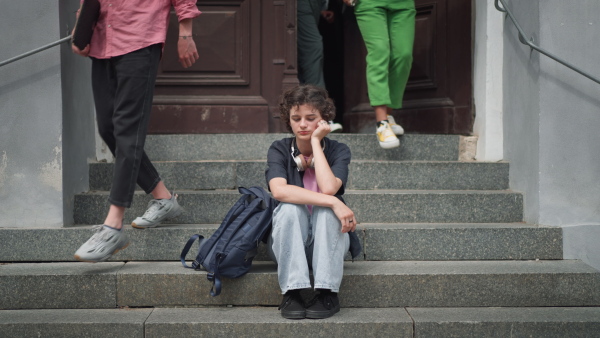 A sad teenager student sitting on stairs in front of school, listening to music. Bullying, discrimination or stress concept.