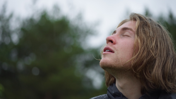 Close-up of a young carefree man standing in forest and enjoying rainy drops at his face.