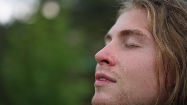 A young carefree handsome young man is feeling free under the rain on a background of green trees.