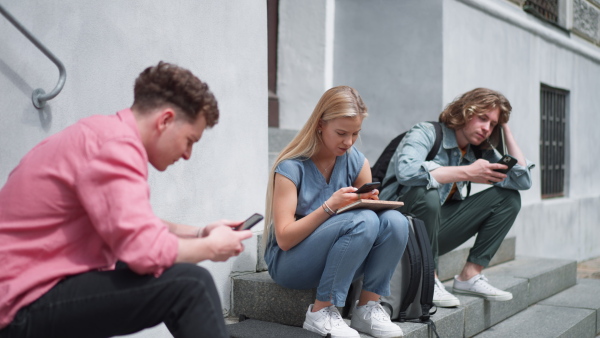 A group of teenagers sitting in outdoor stairs in front of school. Everybody using phone, scrolling. Internet and social media addiction among teenagers concept.