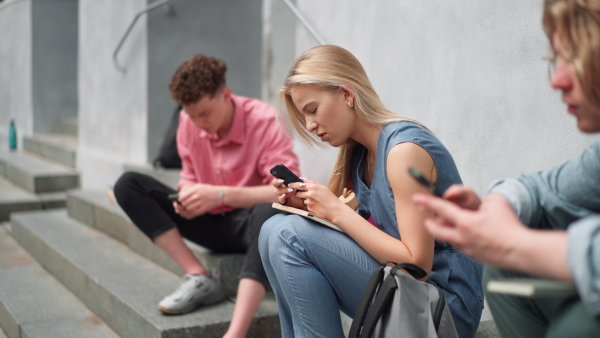 A group of teenagers sitting in outdoor stairs in front of school. Everybody using phone, scrolling. Internet and social media addiction among teenagers concept.