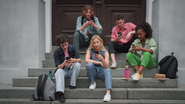 A group of teenagers sitting in outdoor stairs in front of school. Everybody using phone, scrolling. Internet and social media addiction among teenagers concept.