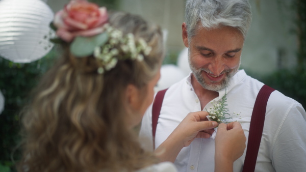 A mature groom recieving decoration on his shirt from bride at wedding reception outside in the backyard.