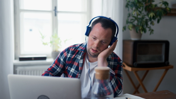 A young man with Down syndrome sitting at desk in office and using laptop, listening to music from headphones.