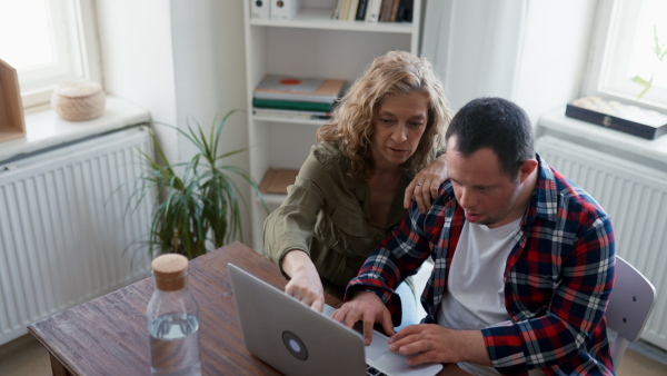 Young man with Down syndrome sitting at desk in office and using laptop, his mother assisting him.