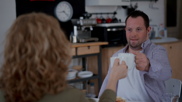 Happy young man with Down syndrome with his mother at home having tea together, clinking with the cups.