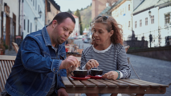 A happy young man with Down syndrome with his mother sitting in cafe outdoors and talking.