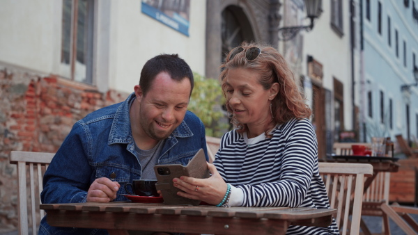 A happy young man with Down syndrome with his mother sitting in cafe outdoors and talking.