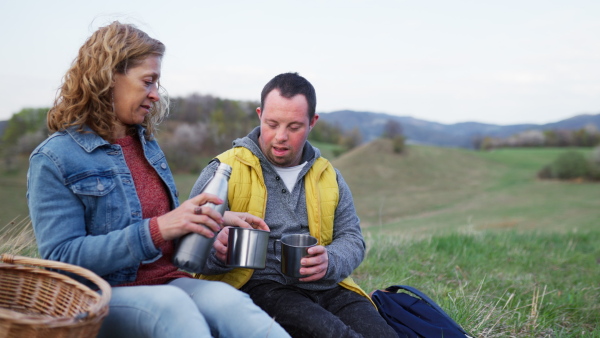 Happy young man with Down syndrome with his mother resting in nature, sitting and drinking tea from a thermos.