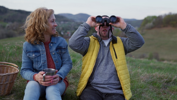 A portrait of happy young man with Down syndrome with his mother resting in nature, sitting and looking at view with binoculars.