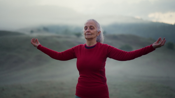 A senior woman doing breathing exercise in nature on early morning with fog and mountains in background.