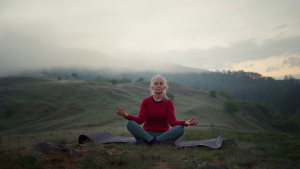 A senior woman doing breathing exercise in nature on early morning with fog and mountains in background.