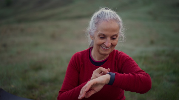 A senior woman jogger setting and looking at sports smartwatch, checking her performance in nature on early morning with fog and mountains in background.