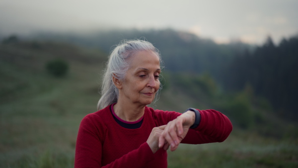 A senior woman jogger setting and looking at sports smartwatch, checking her performance in nature on early morning with fog and mountains in background.